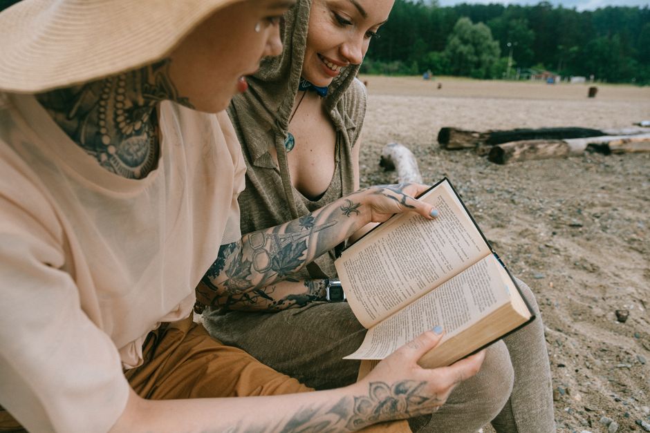 Two tattooed women enjoying a book at a sandy beach, summer leisure activity.